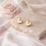 Gold heart-shaped earrings on a soft beige surface with flowers in the background